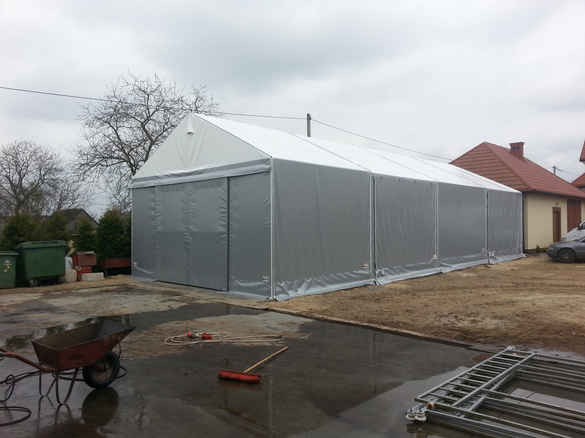 Duży, szary namiot magazynowy z białym dachem, ustawiony na zewnątrz na betonowym podłożu, z widocznymi narzędziami i elementami konstrukcyjnymi w tle.