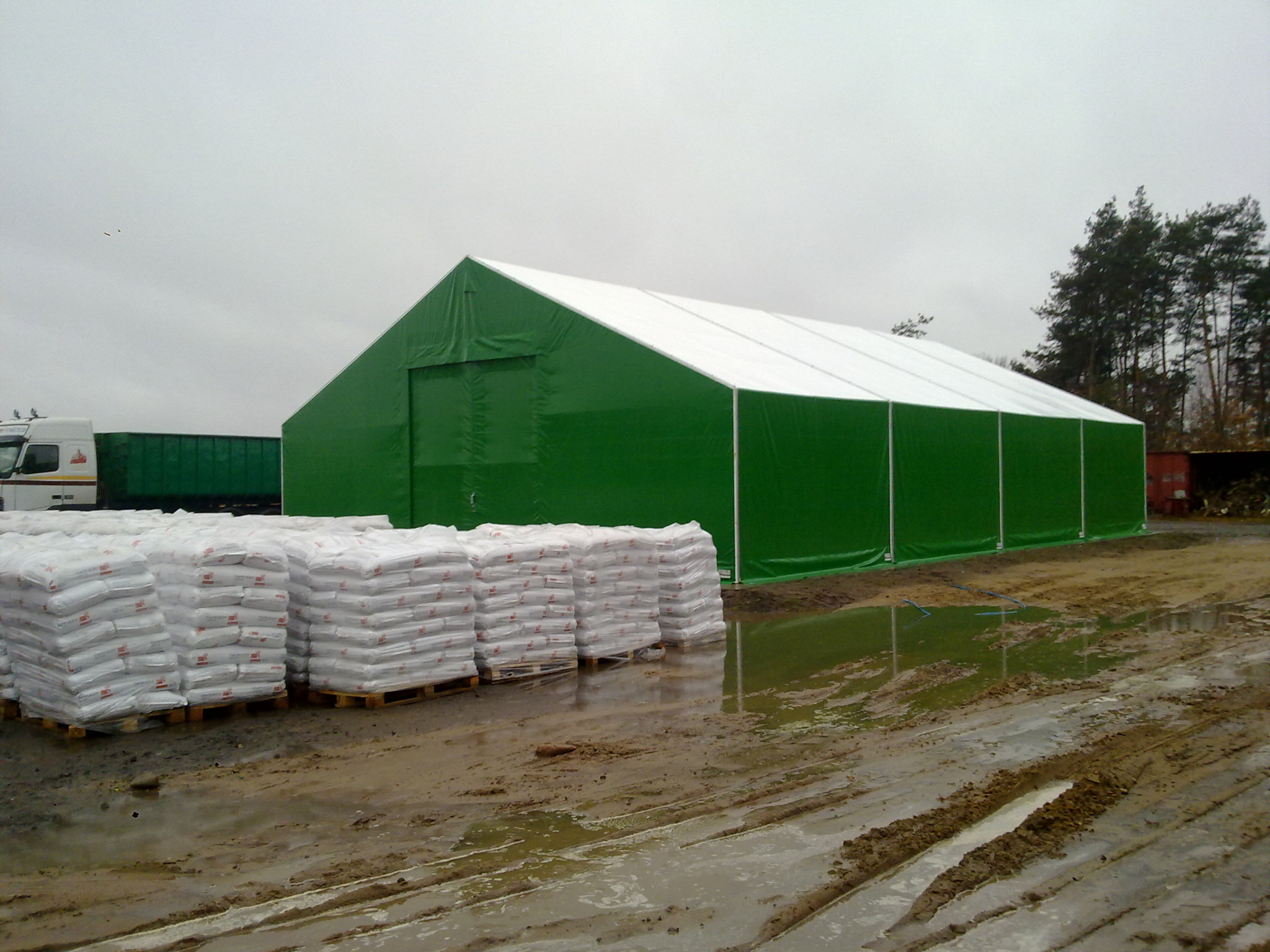 Duży, zielony magazyn namiotowy z białym dachem, palety z workami przed wejściem, błotniste podłoże z odbiciami, ciężarówki w tle, pochmurne niebo.