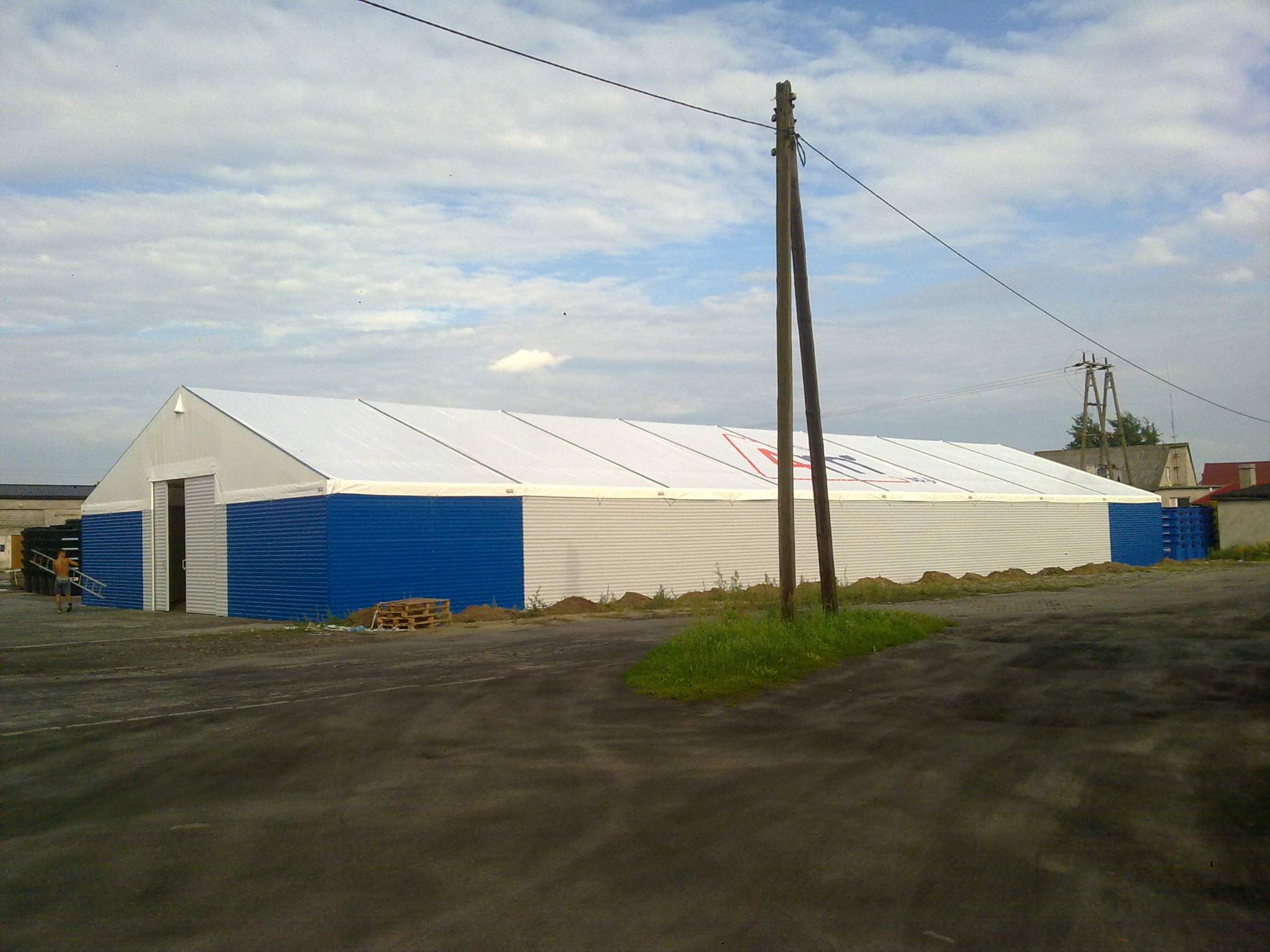 Duży, biało-niebieski magazyn namiotowy z widocznym logo, na tle pochmurnego nieba i asfaltowego placu, z fragmentem osoby niosącej drabinę w tle.