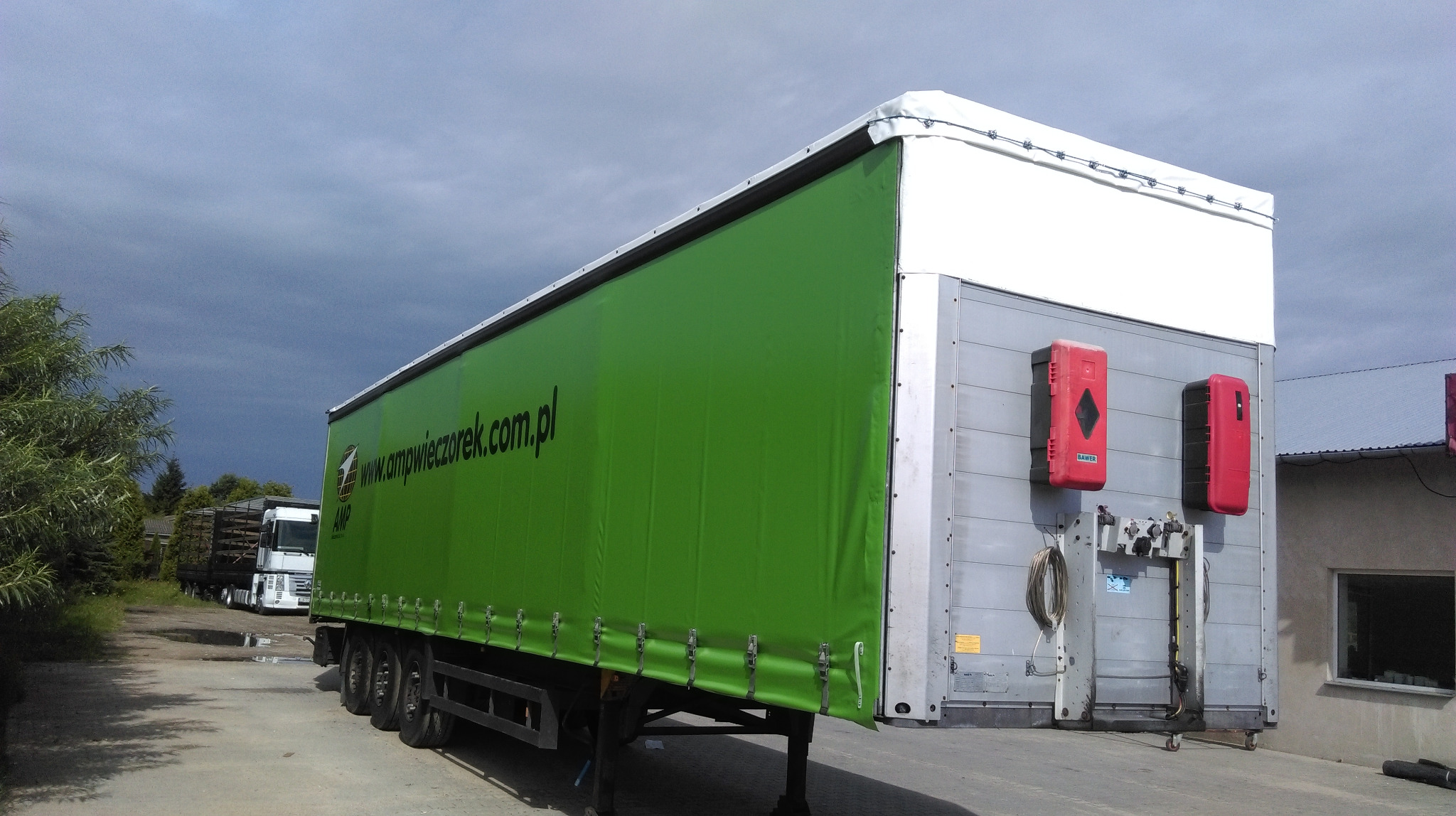 Naczepa ciężarówki z zieloną plandeką i widocznym logo firmy transportowej www.ampwieczorek.com.pl, na tle pochmurnego nieba.