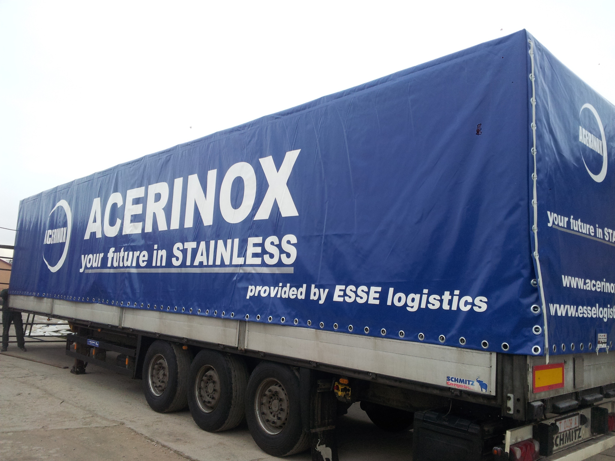 Niebieska plandeka naczepy ciężarówki z białym napisem 'ACERINOX your future in STAINLESS provided by ESSE logistics' oraz logo firmy Acerinox, widok z boku na koła i podwozie.