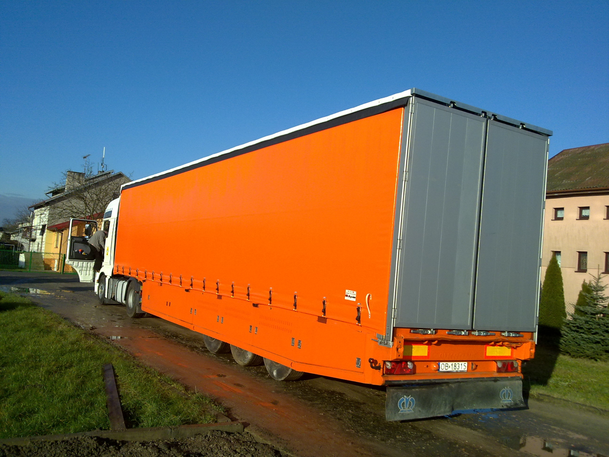 Pomarańczowa naczepa ciężarówki z szarymi drzwiami, widoczna z tyłu, zaparkowana na błotnistej drodze w słoneczny dzień.