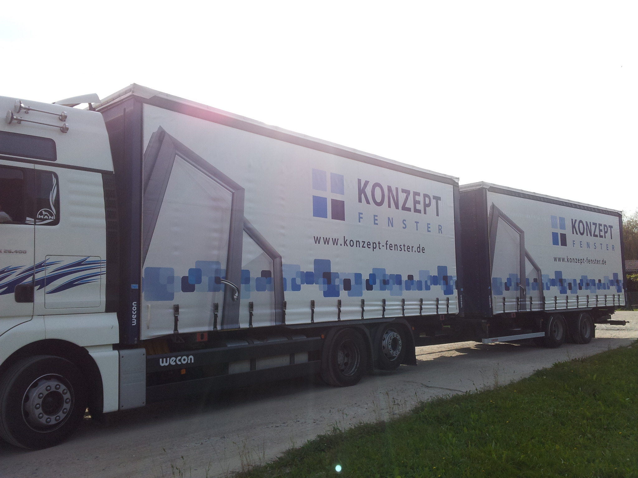 Ciężarówka z naczepą i przyczepą, oklejona reklamą producenta okien z niebieskimi kwadratami i logo 'KONZEPT FENSTER' oraz adresem strony internetowej, zaparkowana na poboczu.