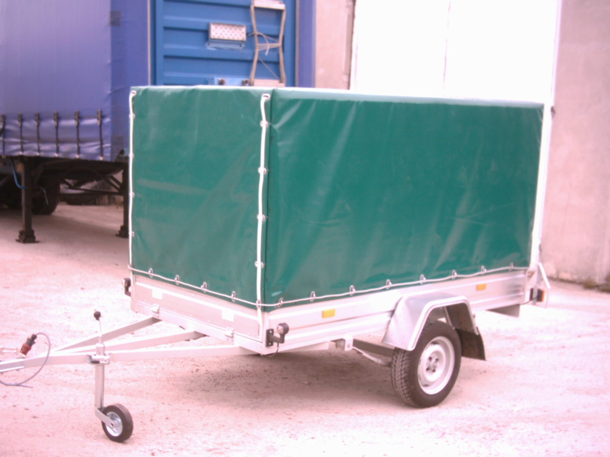 Przyczepa samochodowa z zielonym pokrowcem plandekowym, widok z boku, z tyłu widoczny kontener i inny pojazd.