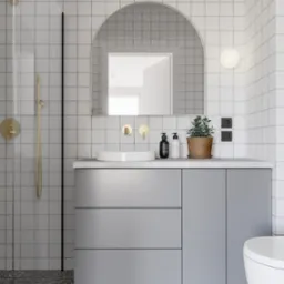 Nowoczesna łazienka z szarymi szafkami, okrągłym lustrem i złotą armaturą. Białe płytki na ścianach i lastryko na podłodze tworzą spójną całość.