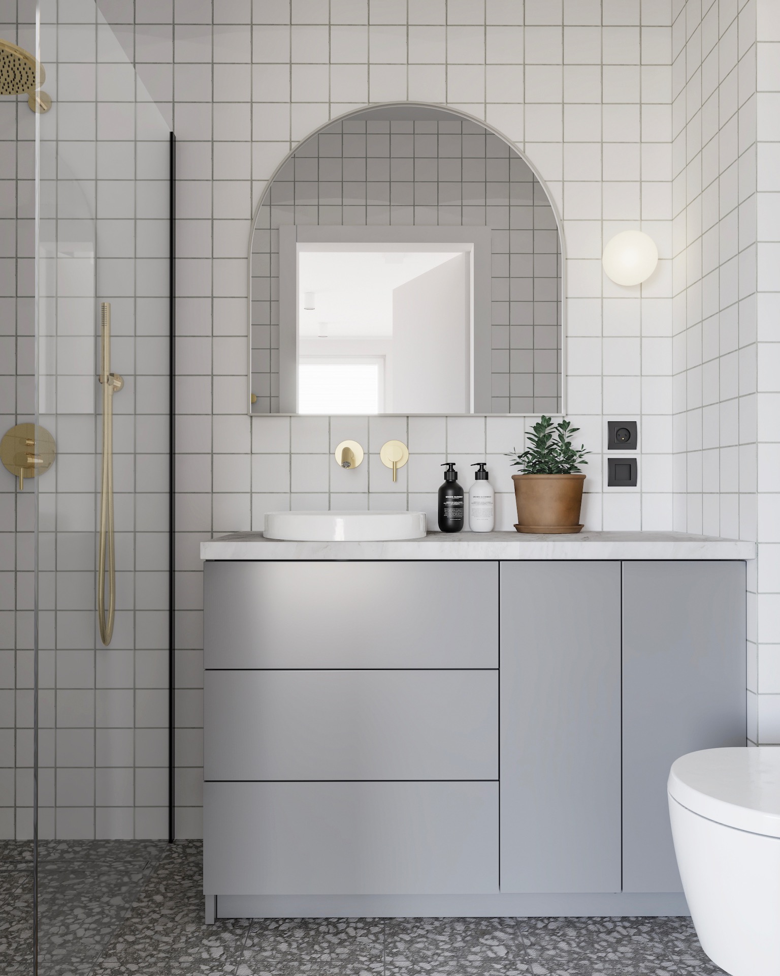 Nowoczesna łazienka z szarymi szafkami, okrągłym lustrem i złotą armaturą. Białe płytki na ścianach i lastryko na podłodze tworzą spójną całość.