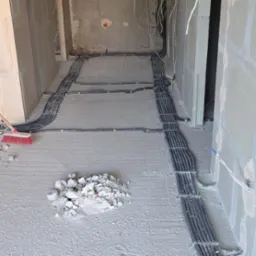 Kable elektryczne w peszlach ułożone na podłodze surowego wnętrza w trakcie prac instalacyjnych, widoczny fragment miotły i gruz.