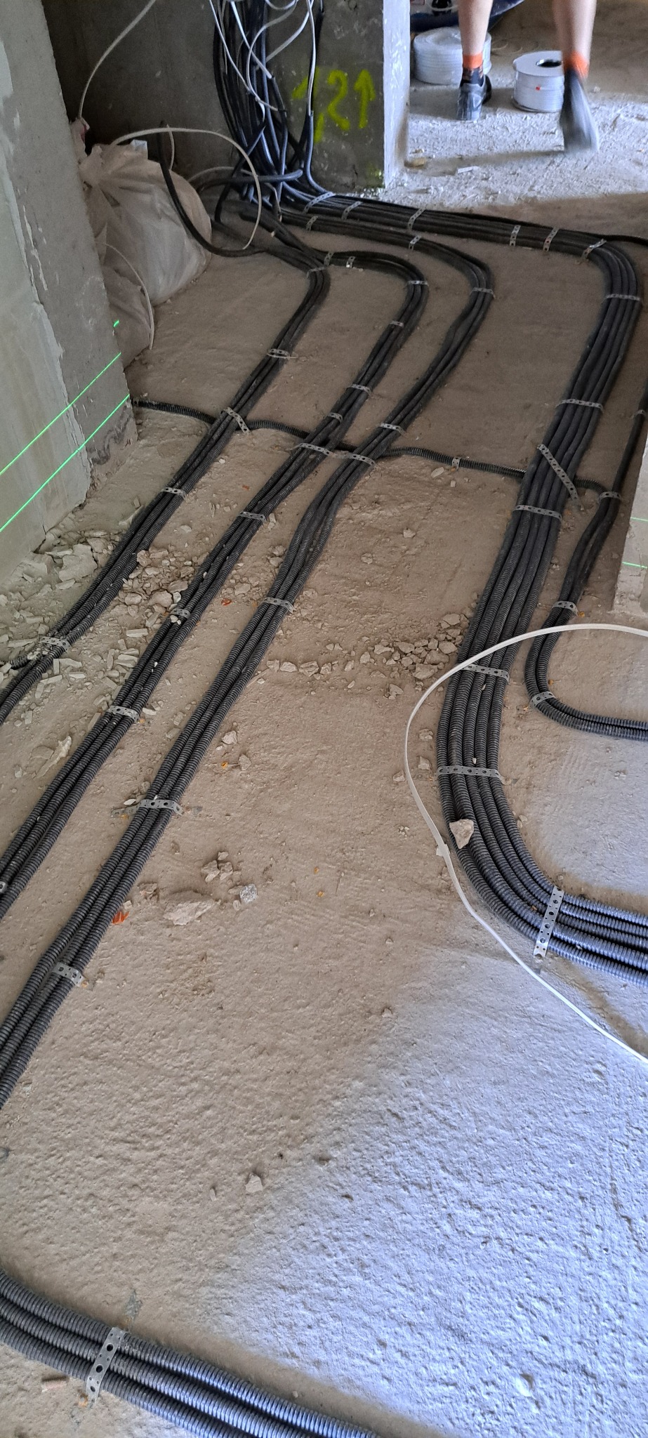 Szare, karbowane rury elektryczne ułożone w równoległych wiązkach na podłodze w surowym stanie, przymocowane metalowymi uchwytami, widoczne ślady cięcia laserem na ścianie.