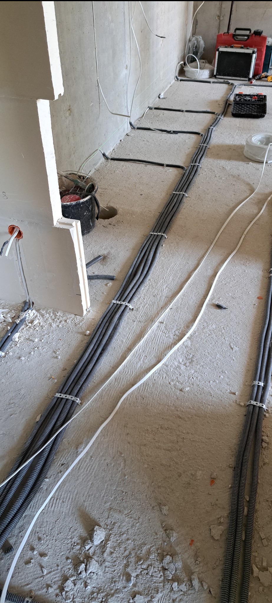 Szare rury instalacji elektrycznej ułożone na surowym betonowym podłożu w trakcie prac wykończeniowych, zabezpieczone metalowymi uchwytami, z widocznymi przewodami i narzędziami w tle.