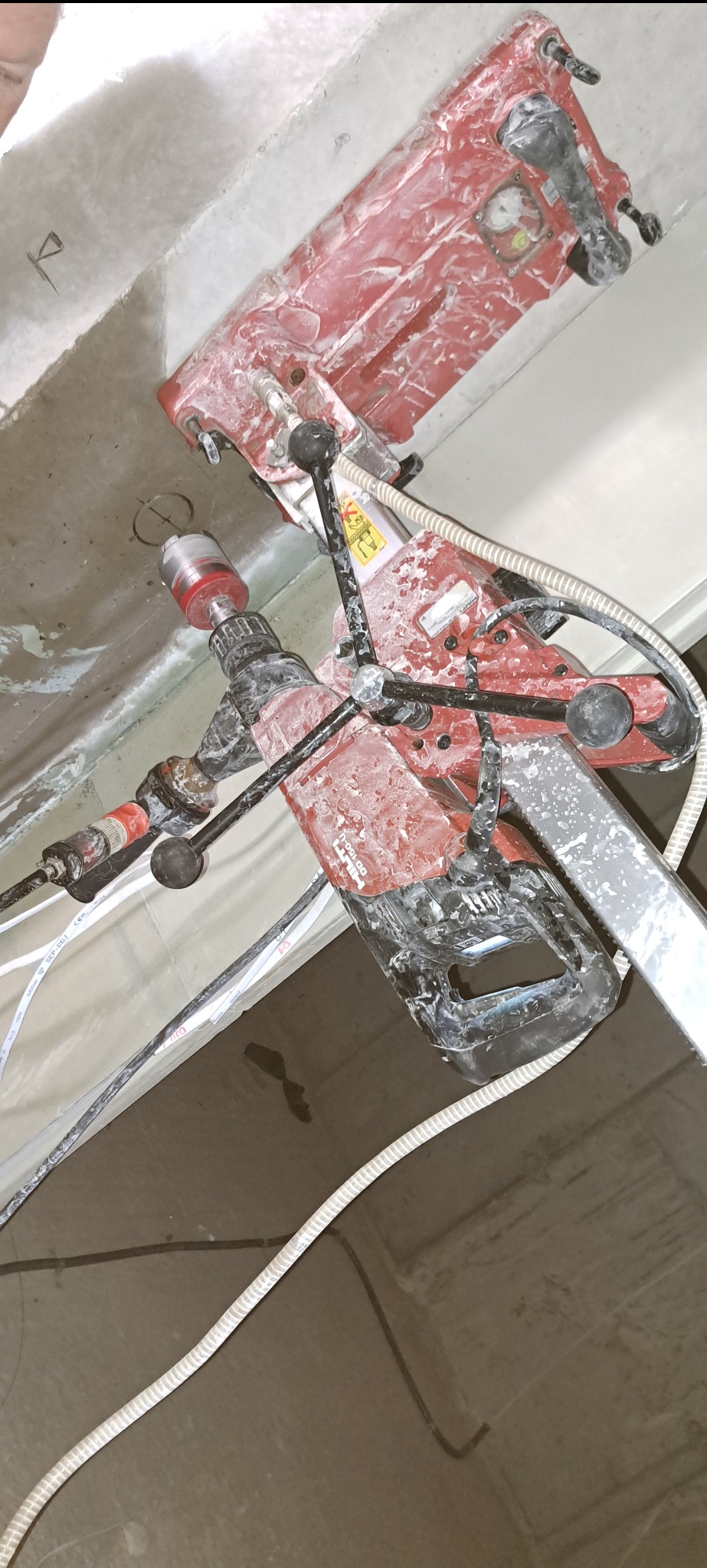 Czerwona wiertnica Hilti DD 150-U, pokryta białym pyłem, wierci otwór w betonowym suficie, widoczne kable i przewody chłodzące.