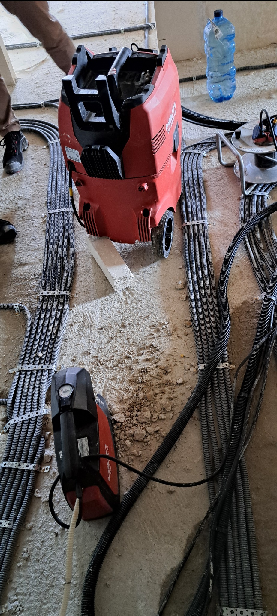 Czerwony odkurzacz przemysłowy Hilti stoi na betonowej podłodze, obok ułożone kable w peszlach, mocowane metalowymi uchwytami. Część kabli ułożona równolegle, inne rozchodzą się w różnych...