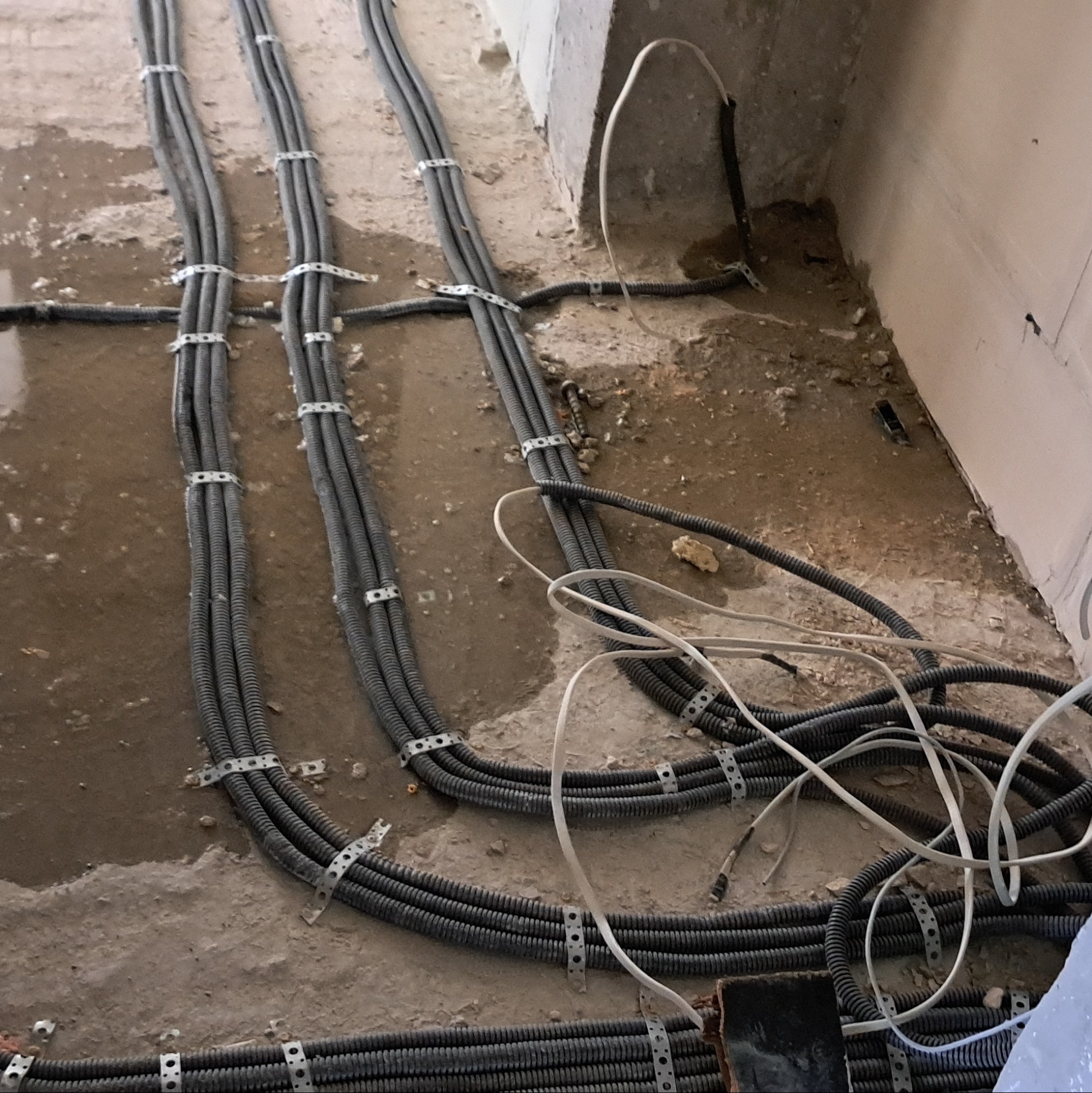 Szare, karbowane rury instalacji elektrycznej, przymocowane metalowymi uchwytami do mokrej, betonowej podłogi w surowym stanie.