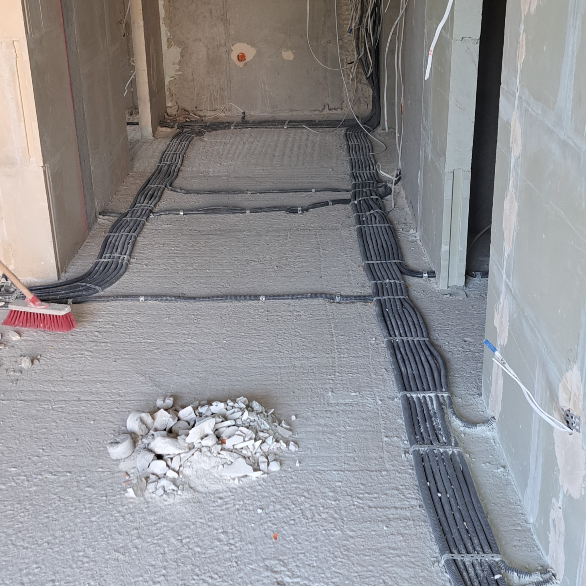 Kable elektryczne w peszlach ułożone na podłodze surowego wnętrza w trakcie prac instalacyjnych, widoczny fragment miotły i gruz.