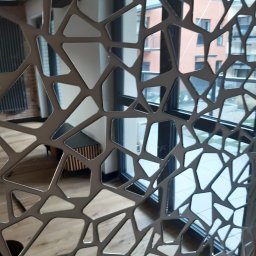 Rk bud - Dekoracyjna ścianka działowa z metalu o ażurowym, geometrycznym wzorze, oddzielająca przestrzeń z drewnianą podłogą i nowoczesną komodą od okna z widokiem na budynek mieszkalny.