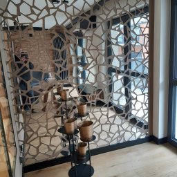 Rk bud - Dekoracyjna ścianka działowa z metalu o nieregularnych kształtach, odbijająca fragment wnętrza z ceglaną ścianą i nowoczesnym grzejnikiem, uzupełniona o stojak z doniczkami w odcieniach brązu.