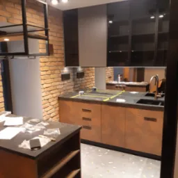 Nowoczesna kuchnia w trakcie montażu: widoczne szafki z ciemnymi frontami i drewnopodobnymi korpusami, ceglana ściana, wyspa kuchenna z blatem roboczym, zabezpieczone folią blaty, oświetlenie...