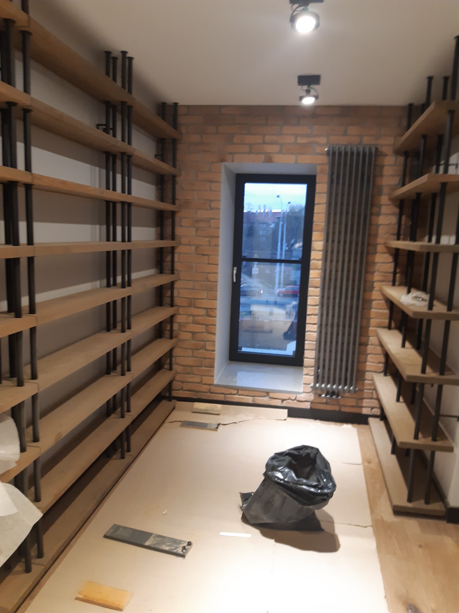 Montaż drewnianych regałów z metalowymi wspornikami w surowym wnętrzu z ceglaną ścianą i zakrytym podłożem, widoczny czarny worek na śmieci.