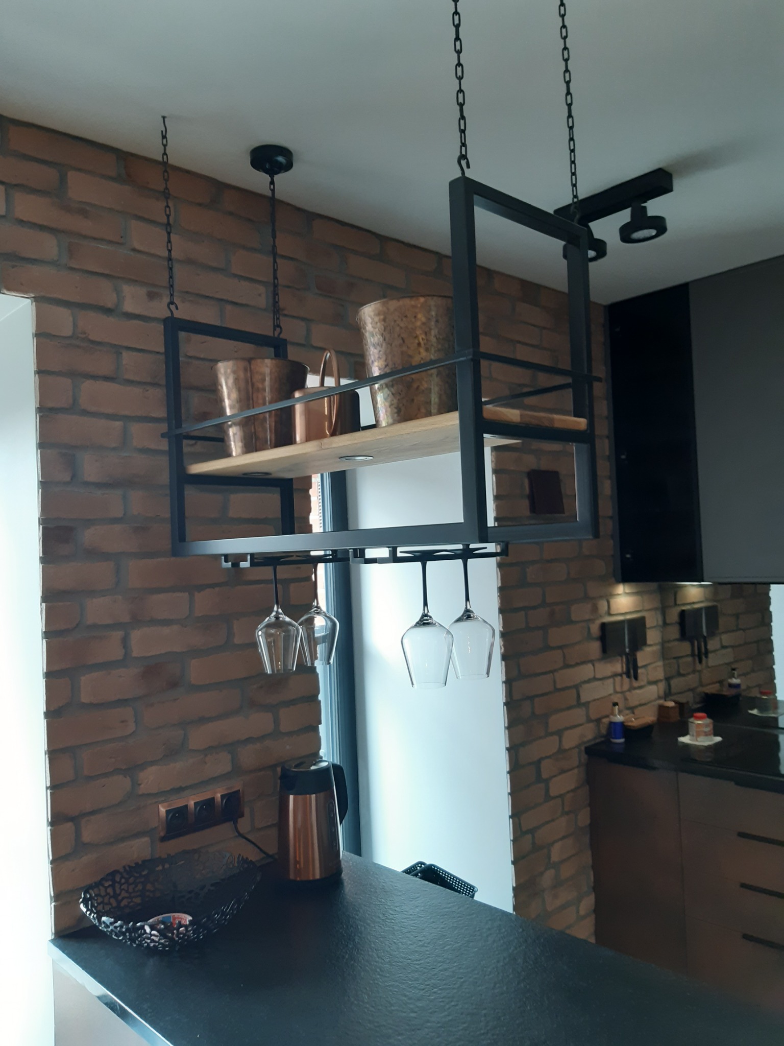 Loftowa kuchnia z ceglaną ścianą, czarnym blatem i metalowym regałem podwieszanym na łańcuchach, eksponującym szklanki do wina i miedziane naczynia.