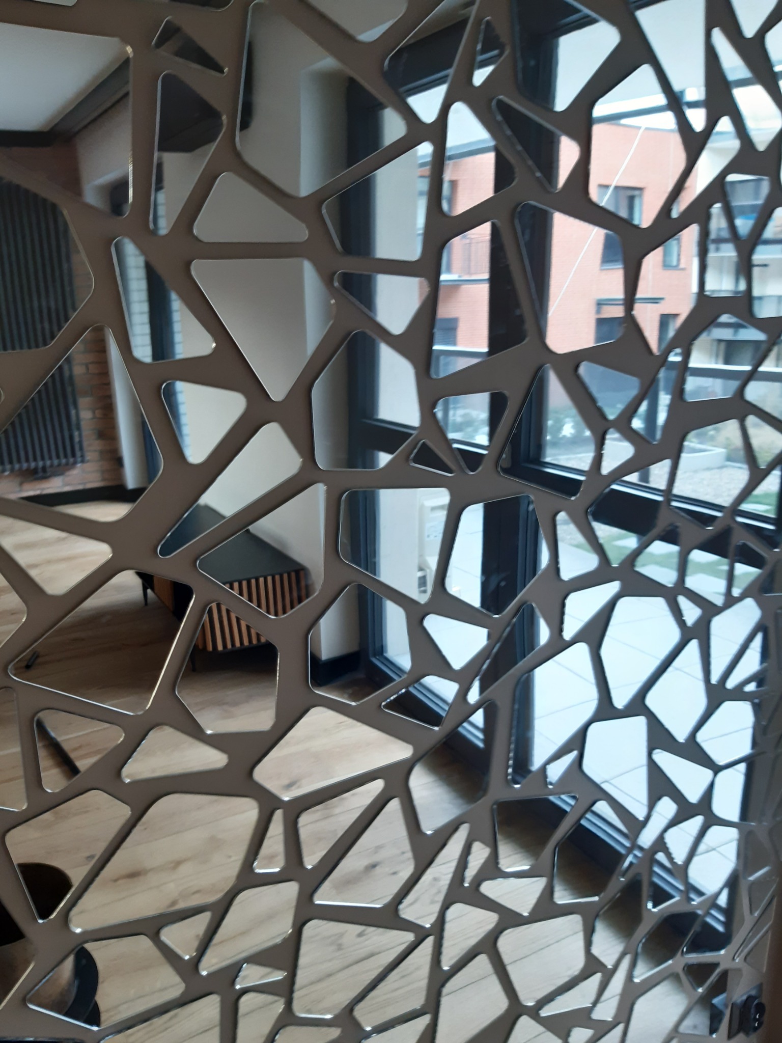 Dekoracyjna ścianka działowa z metalu o ażurowym, geometrycznym wzorze, oddzielająca przestrzeń z drewnianą podłogą i nowoczesną komodą od okna z widokiem na budynek mieszkalny.