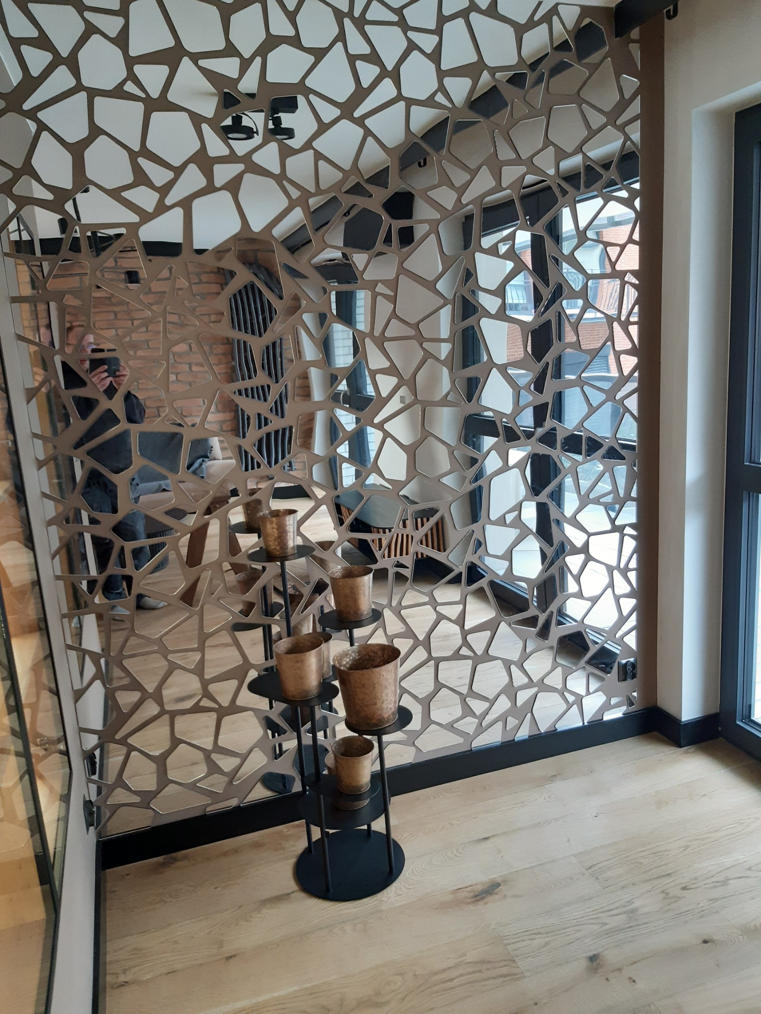 Dekoracyjna ścianka działowa z metalu o nieregularnych kształtach, odbijająca fragment wnętrza z ceglaną ścianą i nowoczesnym grzejnikiem, uzupełniona o stojak z doniczkami w odcieniach brązu.