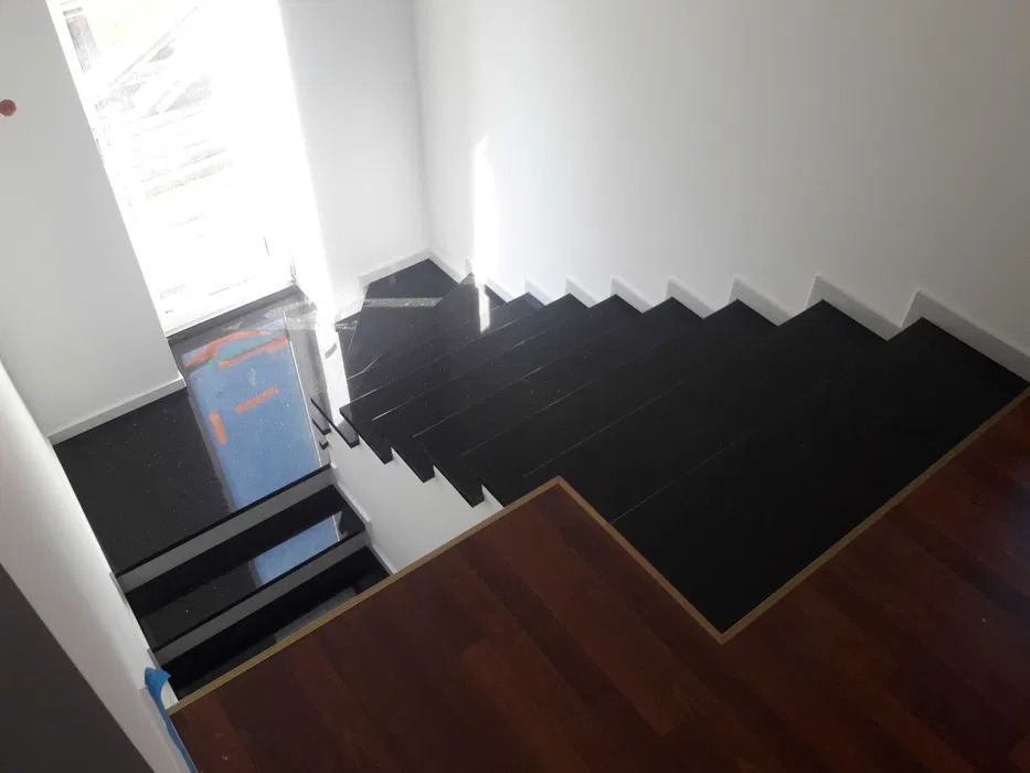 Schody wyłożone czarnym kamieniem, widok z góry, kontrastujące z drewnianą podłogą na dole i białymi ścianami, wpadające światło z okna.