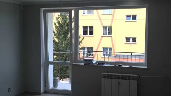 Pokój po malowaniu ścian na szaro, widok na balkon z zielonym drzewem i żółty blok z pomarańczowymi zdobieniami za oknem.
