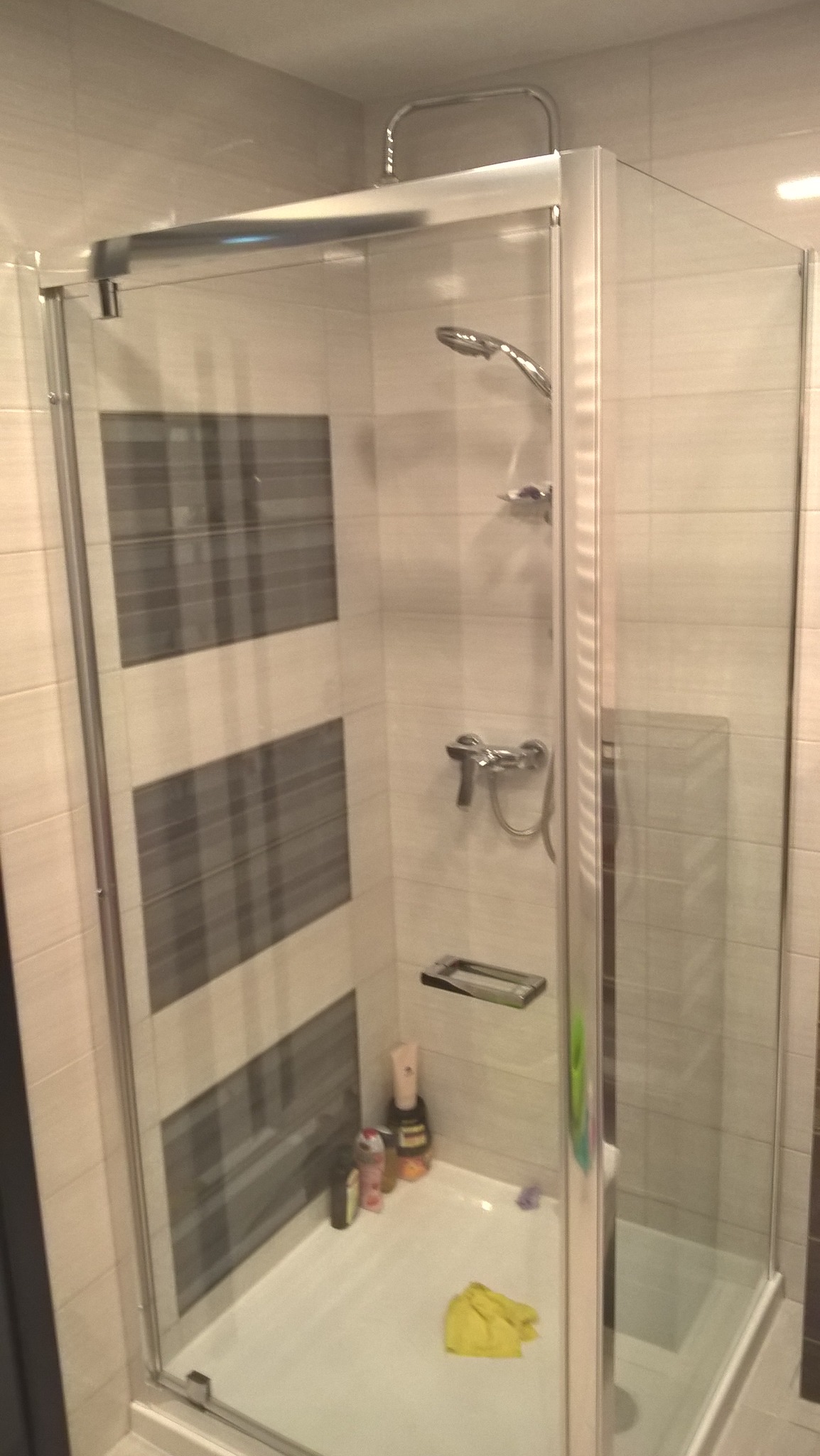 Nowoczesna kabina prysznicowa z chromowanymi profilami i przeszkleniami, widoczne szare pasy na ścianie, bateria, półka na kosmetyki i żółta ścierka w brodziku.