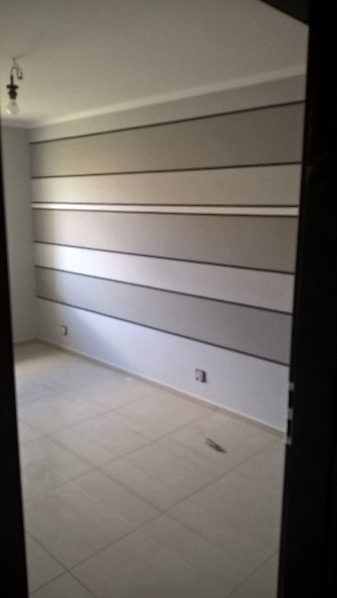 Pomalowane ściany w pokoju z poziomymi pasami w odcieniach szarości i białym sufitem, widok zza drzwi, jasna podłoga z płytek.