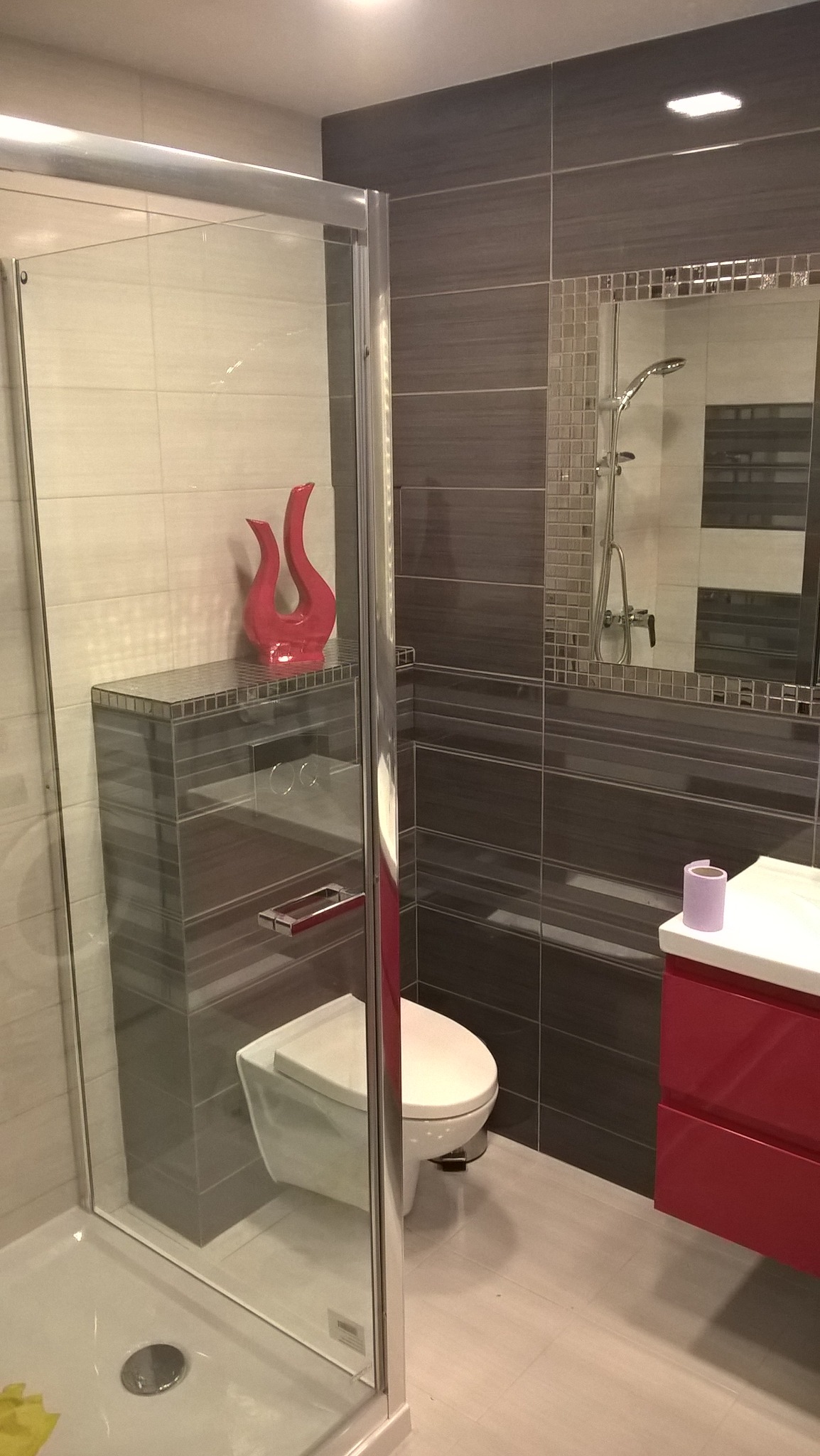 Nowoczesna łazienka z kabiną prysznicową ze srebrnymi ramami, podwieszaną toaletą z szarą obudową, czerwona szafka pod umywalką, lustro z mozaikową ramą i szaro-białe płytki na ścianach.