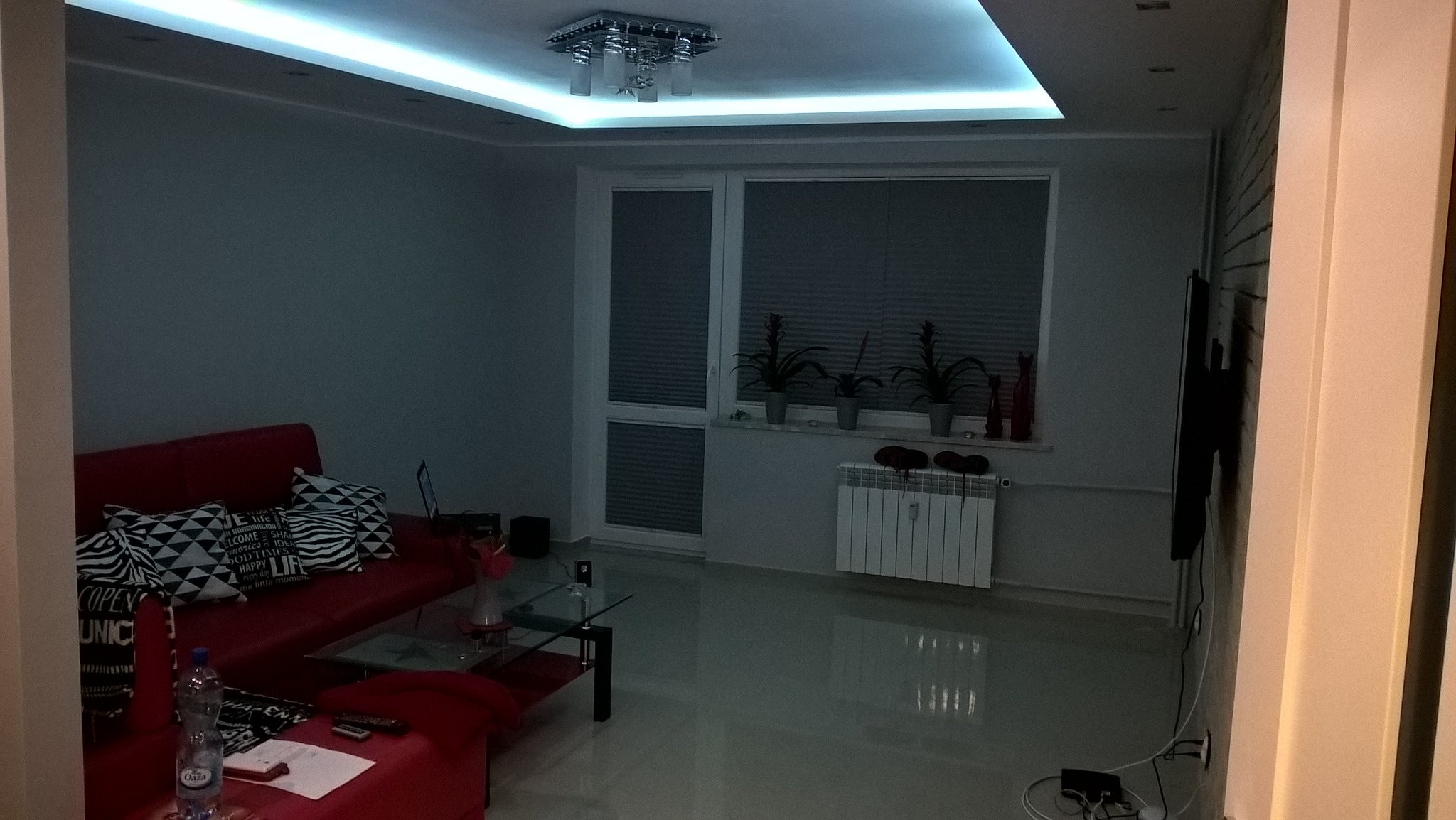 Salon po remoncie: Czerwona sofa z wzorzystymi poduszkami, szklany stolik kawowy, jasna podłoga, okno z żaluzjami, biały grzejnik, oświetlenie sufitowe LED i dekoracje okienne.