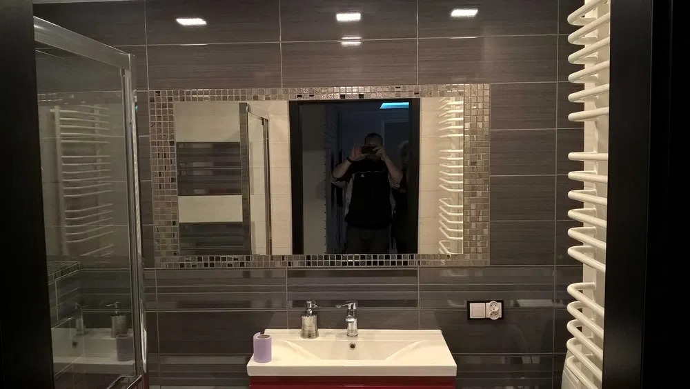 Szara łazienka z kabiną prysznicową, umywalką na czerwonej szafce i lustrem z mozaikową ramą, w którym odbija się osoba robiąca zdjęcie telefonem.