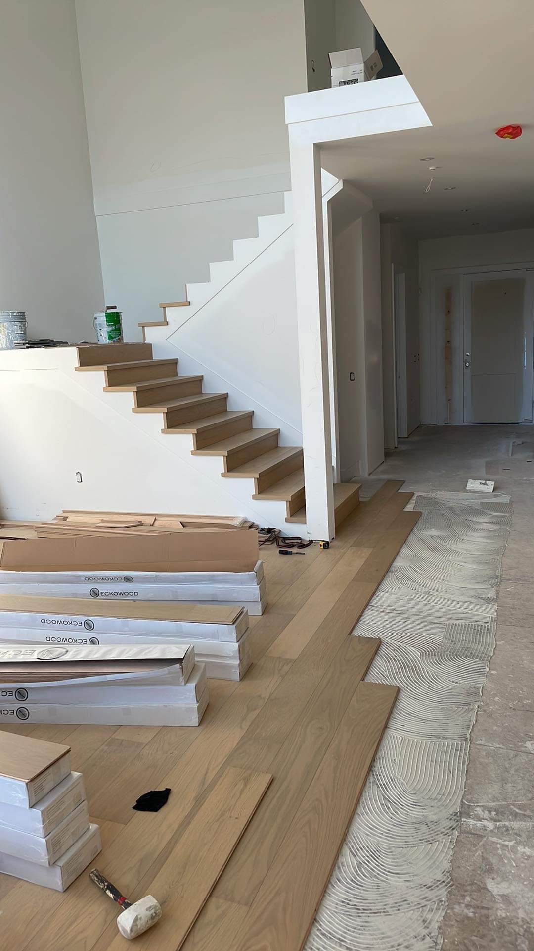 Wnętrze domu w trakcie montażu jasnej drewnianej podłogi, widoczne paczki paneli firmy Eckowood, narzędzia, schody z drewnianymi stopniami i rozprowadzony klej na posadzce.