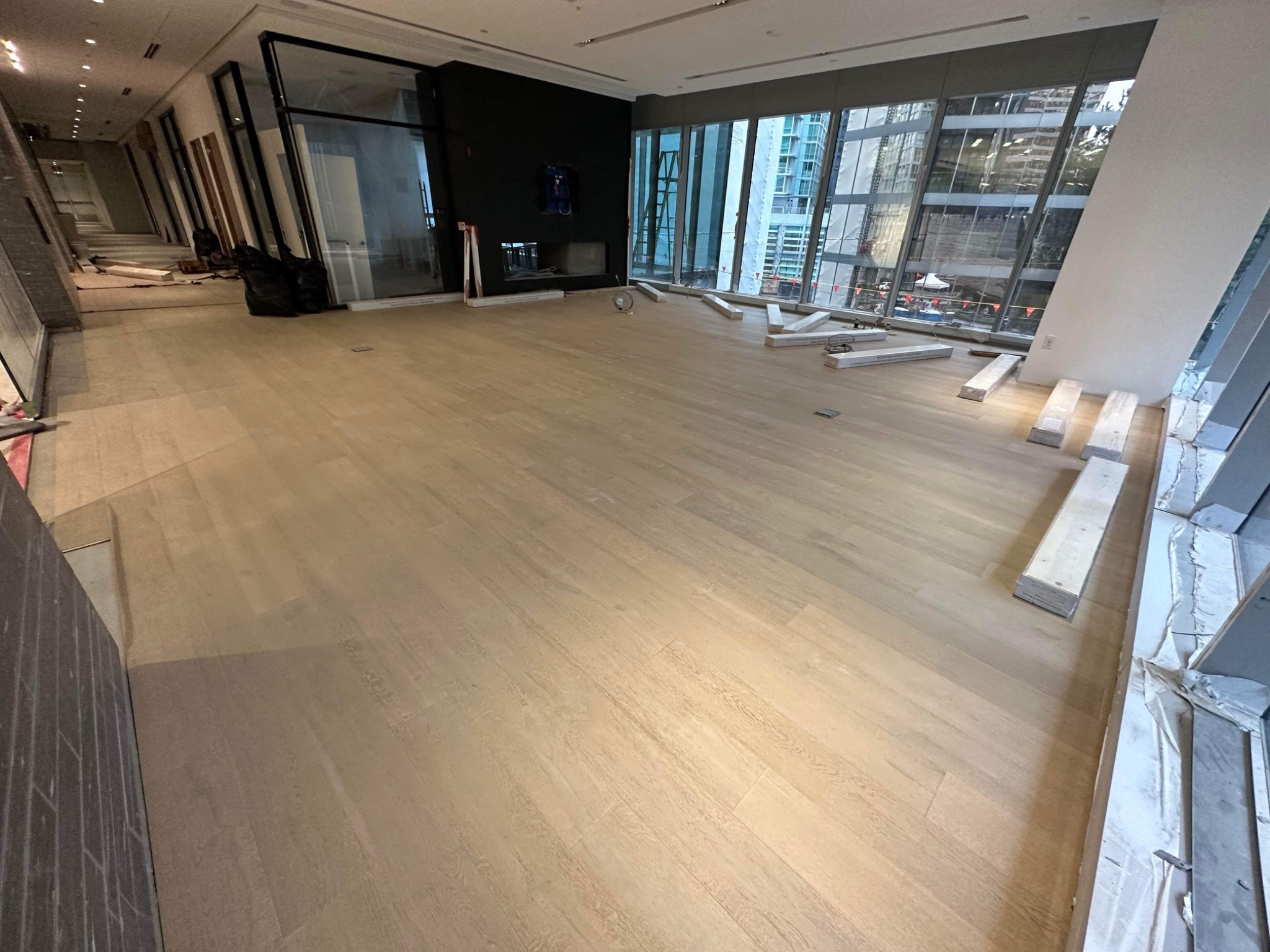 Jasna, nowo ułożona podłoga w przestronnym wnętrzu z dużymi oknami, widoczne paczki paneli czekające na montaż wzdłuż ściany, czarny kominek w tle.
