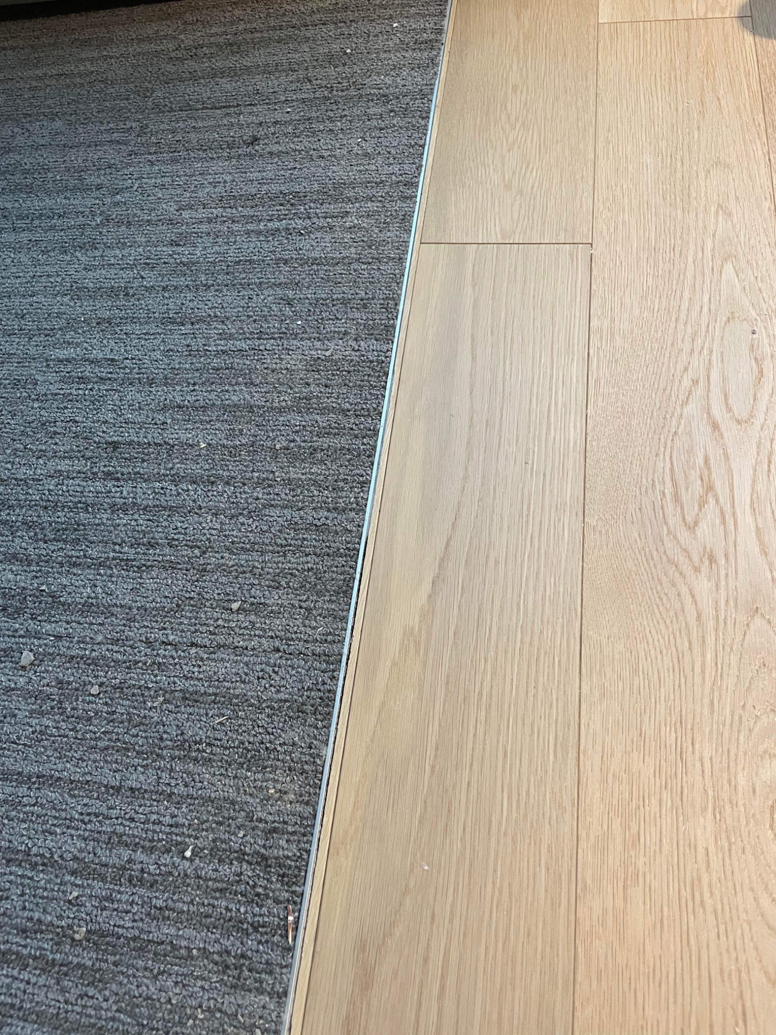 Połączenie jasnych paneli podłogowych z szarym dywanem, listwa przejściowa widoczna.