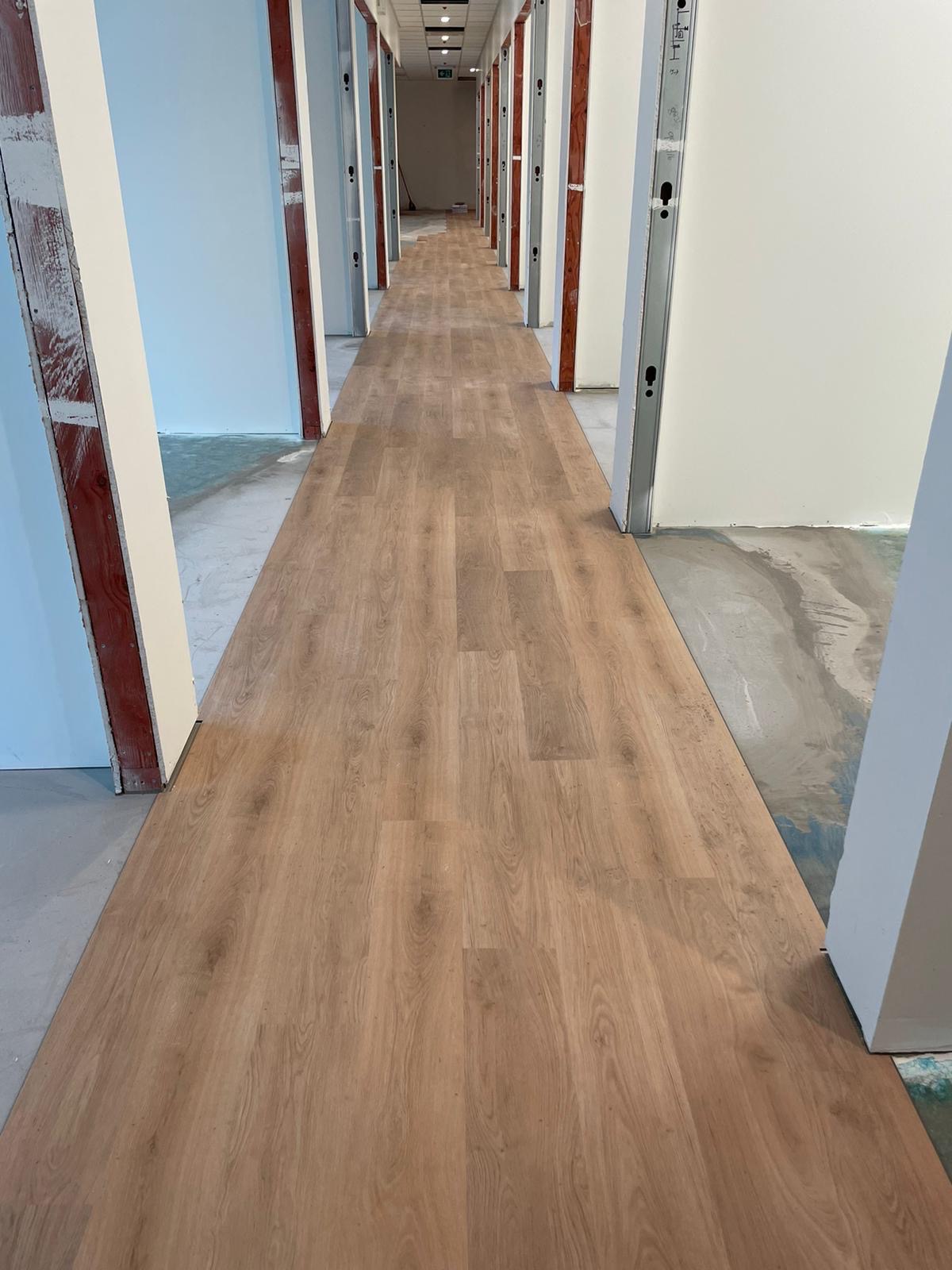 Długi korytarz z nowo położoną, jasną podłogą imitującą drewno, widoczne surowe ściany i futryny w trakcie wykończenia.