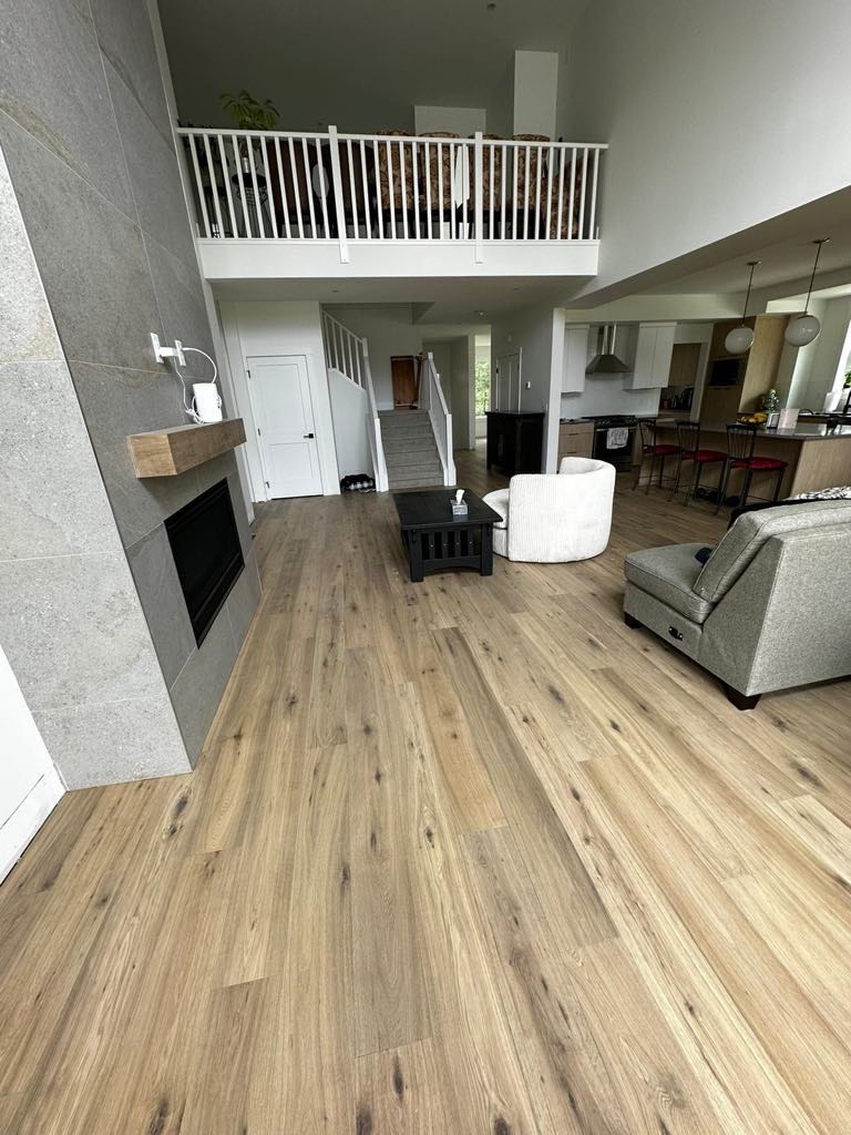 Jasna, drewnopodobna podłoga w przestronnym salonie z kominkiem i antresolą, widok na otwartą kuchnię.