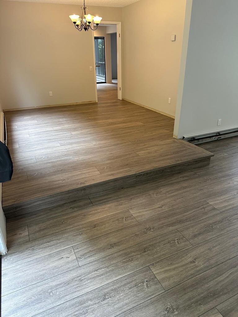 Jasnoszara podłoga z paneli imitujących drewno, z widocznym progiem i przejściem do kolejnego pomieszczenia, oświetlona przez wiszącą lampę.