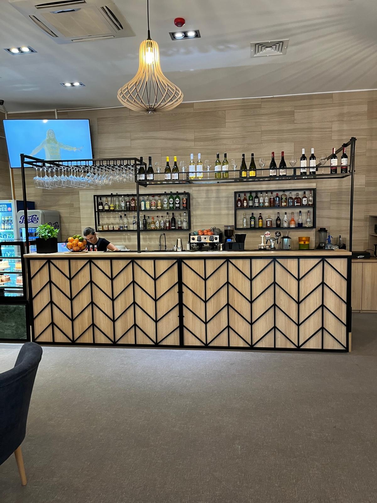 Bar w restauracji z metalową konstrukcją w kształcie strzałek na drewnianych panelach, wiszące kieliszki i półki z butelkami alkoholu, oświetlenie wiszące, osoba za barem i telewizor w tle.