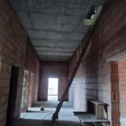 Wnętrze surowego, murowanego domu w trakcie budowy, widok korytarza z ceglanymi ścianami, betonowym stropem z otworem, drewnianym stołem warsztatowym i drabiną opartą o ścianę.