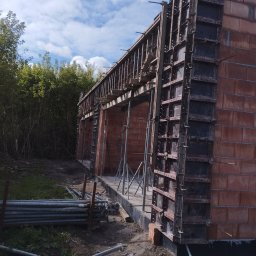 R-C BUD Robert Czuj - Wznoszenie ścian z czerwonej cegły z użyciem szalunków systemowych, podpory stalowe i leżące rury budowlane na placu budowy, widok na błękitne niebo.