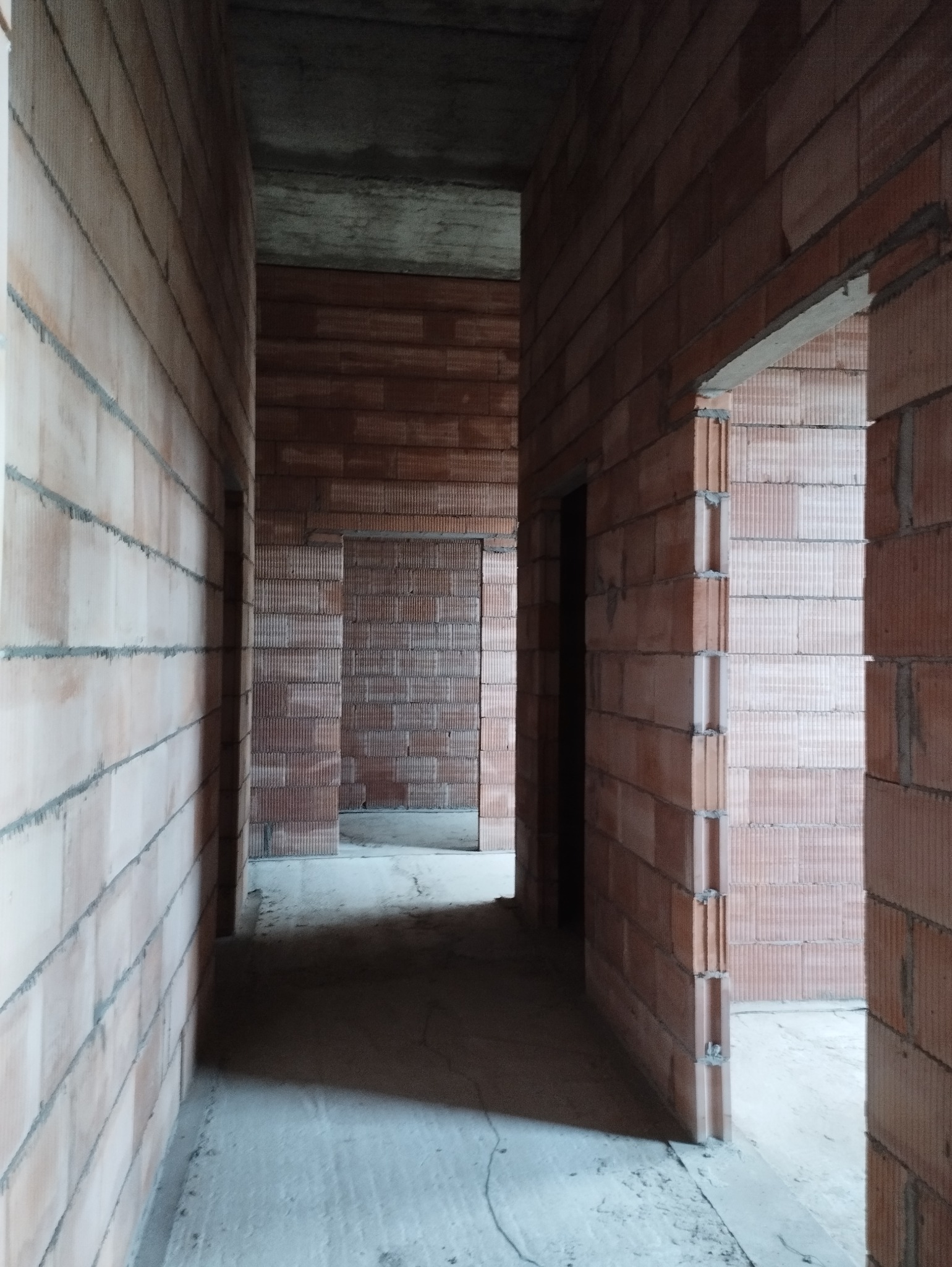 Wnętrze surowego domu w trakcie budowy, widoczne ściany z czerwonej cegły ceramicznej, surowy betonowy strop i otwory drzwiowe bez ościeżnic.