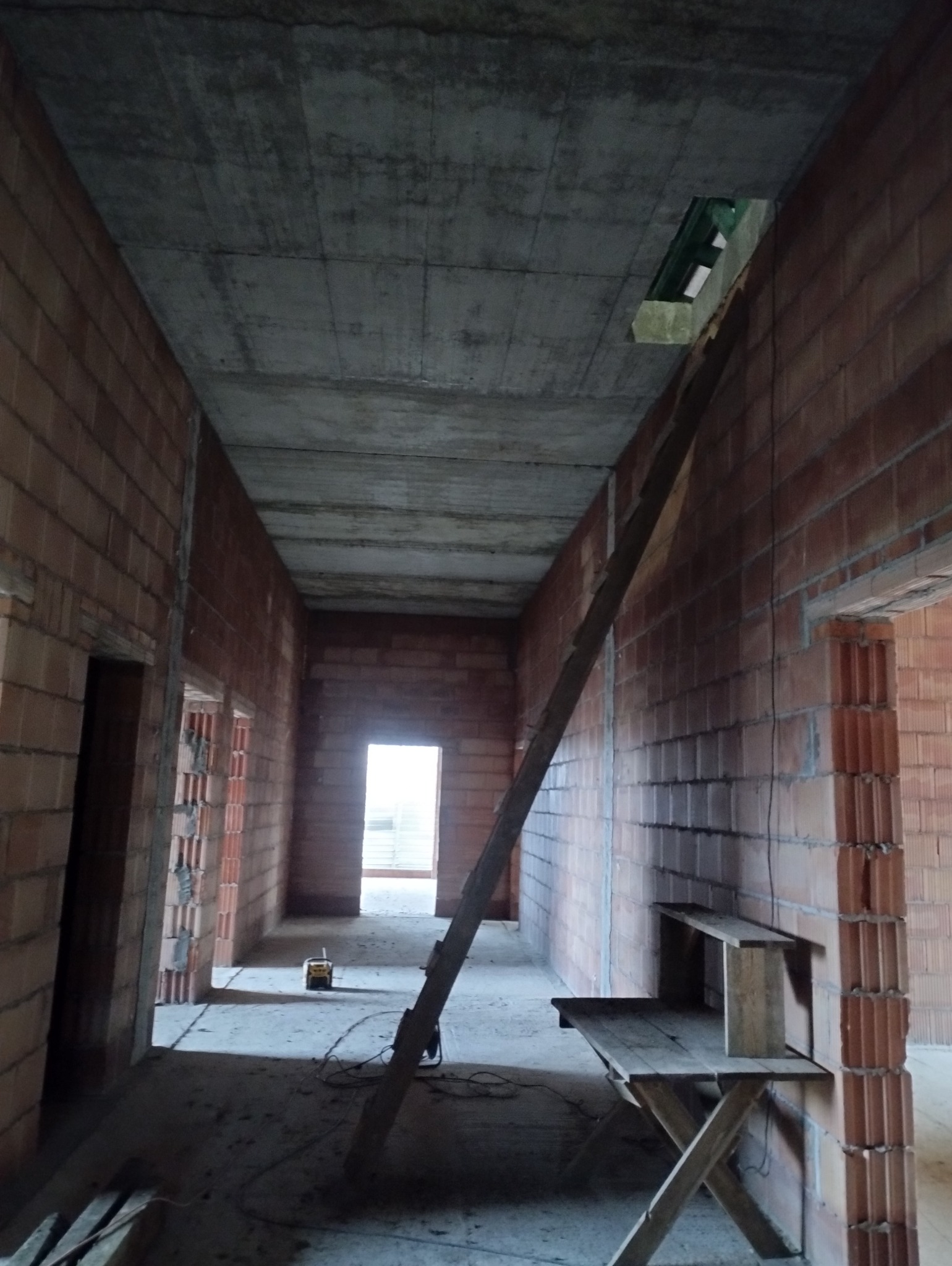 Wnętrze surowego, murowanego domu w trakcie budowy, widok korytarza z ceglanymi ścianami, betonowym stropem z otworem, drewnianym stołem warsztatowym i drabiną opartą o ścianę.