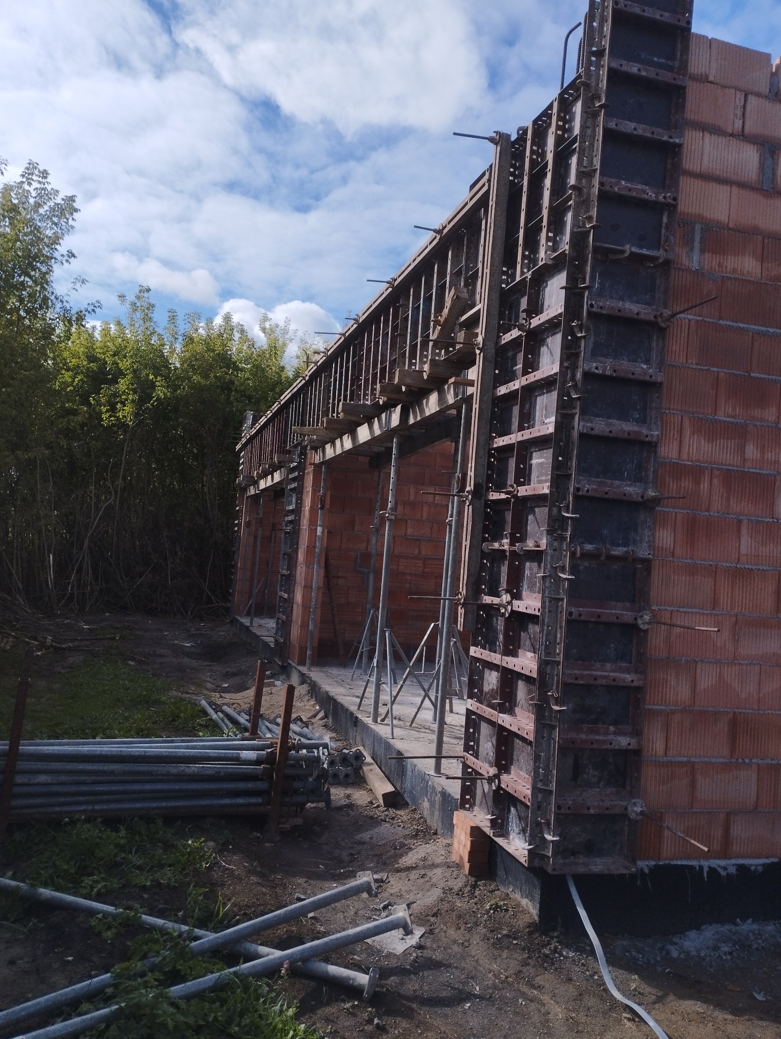 Wznoszenie ścian z czerwonej cegły z użyciem szalunków systemowych, podpory stalowe i leżące rury budowlane na placu budowy, widok na błękitne niebo.