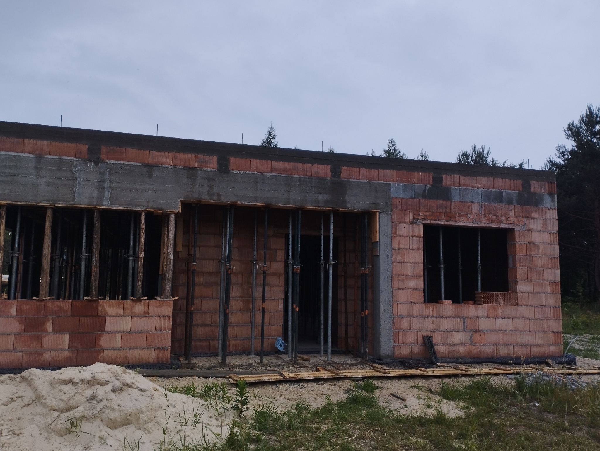 Surowy stan domu jednorodzinnego w trakcie budowy: widoczne ściany z czerwonej cegły, betonowe nadproża i stalowe podpory szalunkowe przy otworach okiennych i drzwiowych, zwały piasku przed budynkiem.