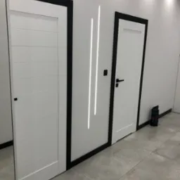 Nowoczesne, białe drzwi z czarnymi ościeżnicami i minimalistycznym oświetleniem liniowym LED na białej ścianie, szara posadzka z płytek.