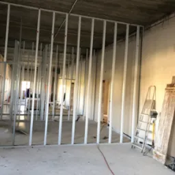 Montaż metalowego stelażu pod ścianę działową z płyty gipsowo-kartonowej w surowym wnętrzu, widoczna drabina i przewód elektryczny na podłodze.