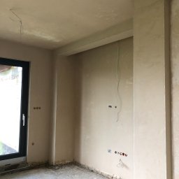 M.HOUSE - Wnętrze w trakcie prac wykończeniowych: ściany pokryte świeżym tynkiem, widoczne gniazdka elektryczne i wystające przewody, surowa podłoga.