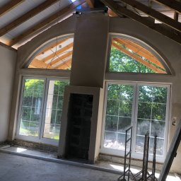 M.HOUSE - Wnętrze w trakcie prac wykończeniowych: surowy komin, duże okna łukowe z widokiem na zieleń, widoczne elementy konstrukcji dachu z drewnianych belek, podłoga w stanie surowym, widoczne podpory...