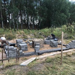 M.HOUSE - Budowa fundamentów domu z bloczków betonowych na piaszczystym terenie, widoczni dwaj pracownicy w trakcie murowania, w tle las.