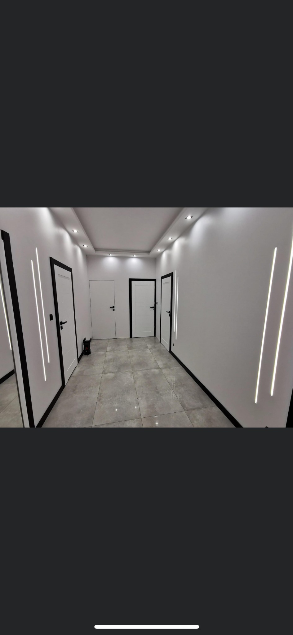 Nowoczesny korytarz z białymi ścianami, szarą podłogą z płytek, białymi drzwiami z czarnymi ościeżnicami i ledowym oświetleniem liniowym.